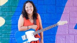 A photo of a woman holding a guitar in front of a colorful background